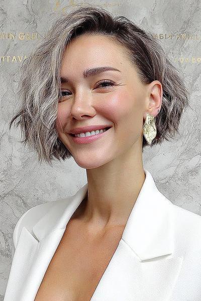 Een vrouw met een vlot, kort kapsel in een asymmetrische bob, met golvend lichtbruin haar en een zijscheiding.