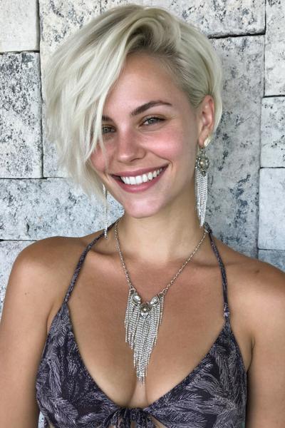 Een vrouw met kort, asymmetrisch blond haar dat aan één kant langer is, glimlacht naar de camera.