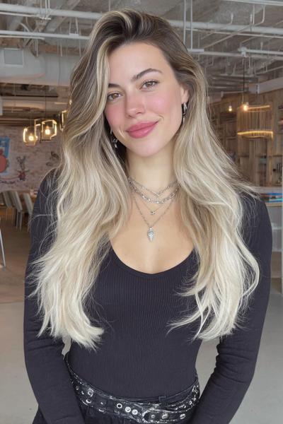 Een vrouw met lang, golvend blond haar met donkere uitgroei en een middenscheiding, gekleed in een zwarte top.