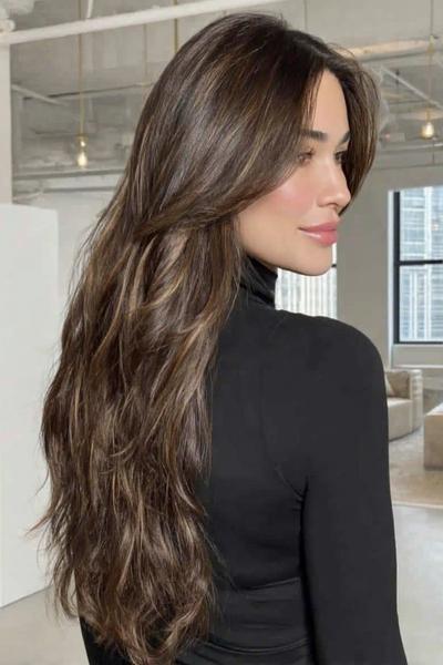 Een vrouw met lang, golvend bruin haar en subtiele highlights, gestyled met een middenscheiding en zachte lagen.
