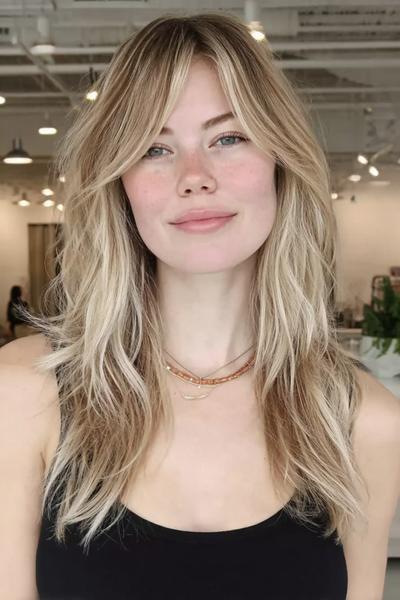 Een vrouw met lang, golvend blond haar en gezichtsomlijstende lagen, geknipt in een natuurlijke, nonchalante stijl.