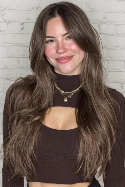 Een jonge vrouw met lang, golvend bruin haar en gezicht omlijstende lagen, gekleed in een donkerbruin shirt.
