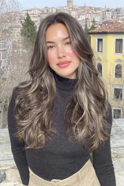 Een vrouw met lang, golvend haar en subtiele highlights poseert buiten in een stadse omgeving.