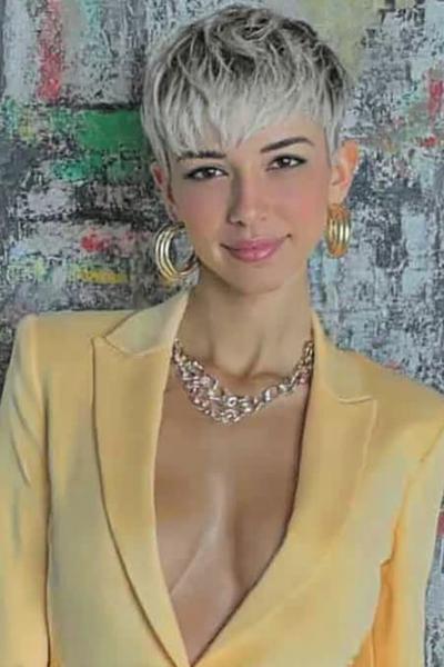 Een vrouw met een kort, gelaagd kapsel met blonde highlights en een pony, gekleed in een gele blazer.