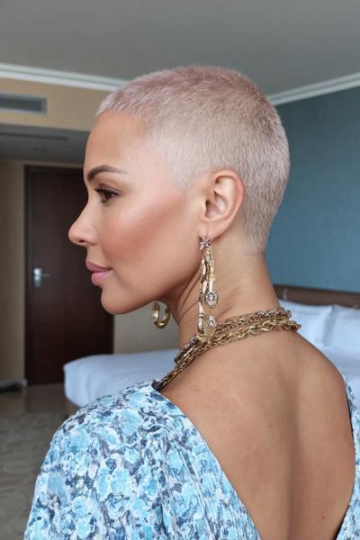 Een vrouw met een kort, geplatinumeerd buzzcut kapsel, draagt gouden sieraden en een lichtblauwe top.