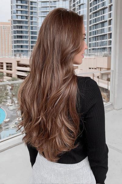 Een vrouw met lang, golvend bruin haar dat los over haar rug valt, gekleed in een zwarte trui en een lichtgrijze rok.