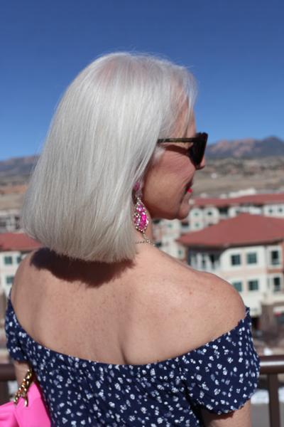 Een oudere dame met recht, schouderlang grijs haar in een klassieke bob kapsel, draagt een off-shoulder jurk en zonnebril.