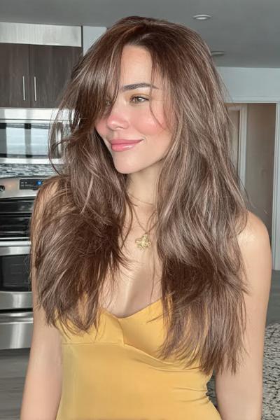 Een vrouw met lang, gelaagd bruin haar met losse golven en een zijscheiding.