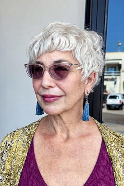 Een oudere vrouw met een kort, golvend en wit kapsel, draagt een bril en blauwe oorbellen.