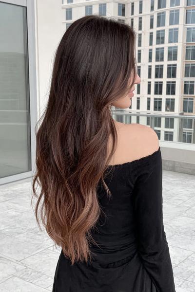 Een vrouw met lang, golvend bruin haar draagt een off-shoulder zwart shirt en staat op een balkon met een stedelijke achtergrond.