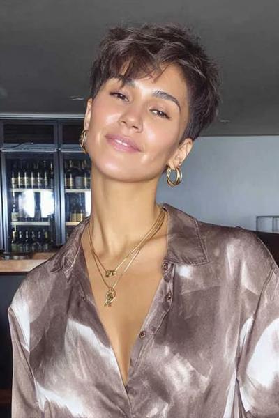 Een vrouw met een kort, golvend kapsel en een zijscheiding, gekleed in een glanzende blouse en gouden sieraden.