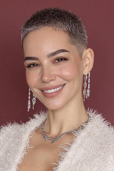 Een vrouw met een zeer kort geschoren kapsel en een glimlach, draagt zilveren oorbellen en een witte, pluizige trui.