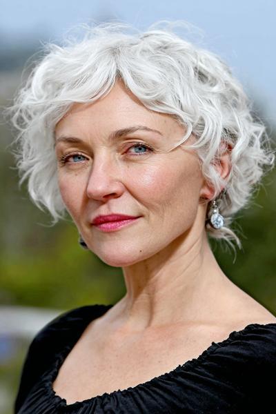 Een oudere dame met kort, wit, krullend haar en een speels, vlot kapsel.