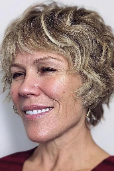 Een vrouw met kort, golvend blond haar in een speels en volumineus kapsel, geschikt voor dames van 60 plus.